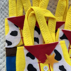 Puede incluir: Primer plano de varias bolsas de mano de fieltro amarillo con asas largas. Las bolsas presentan un dise&ntilde;o inspirado en un personaje de vaquero, con un pa&ntilde;uelo rojo, un chaleco blanco con manchas negras y una placa de sheriff. Las bolsas tienen detalles en marr&oacute;n, azul y amarillo.