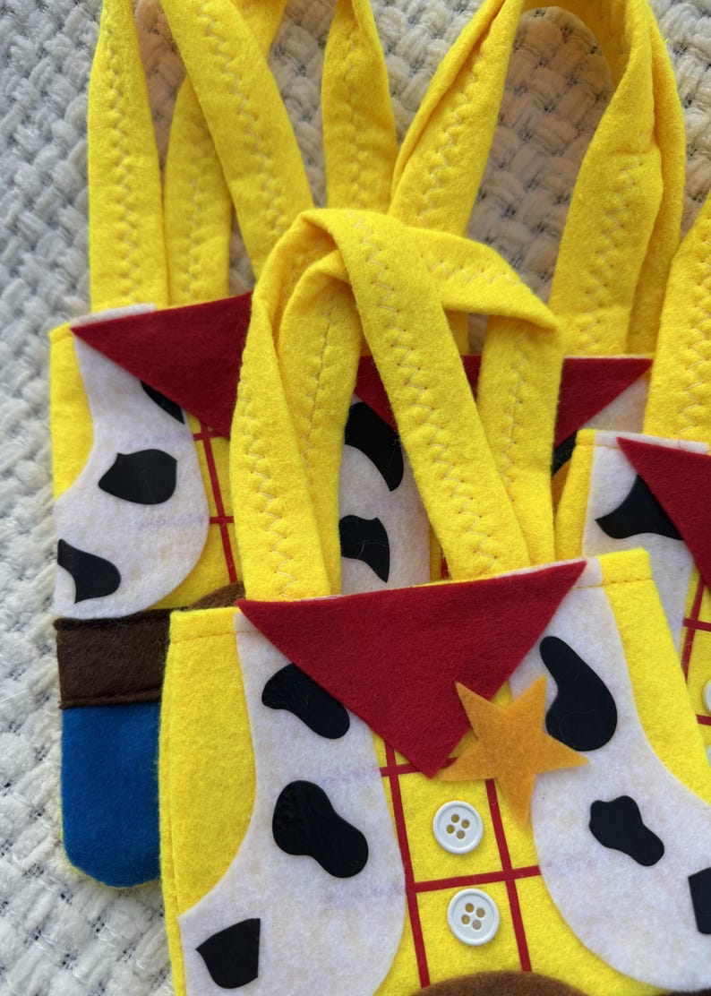 Puede incluir: Primer plano de varias bolsas de mano de fieltro amarillo con asas largas. Las bolsas presentan un dise&ntilde;o inspirado en un personaje de vaquero, con un pa&ntilde;uelo rojo, un chaleco blanco con manchas negras y una placa de sheriff. Las bolsas tienen detalles en marr&oacute;n, azul y amarillo.