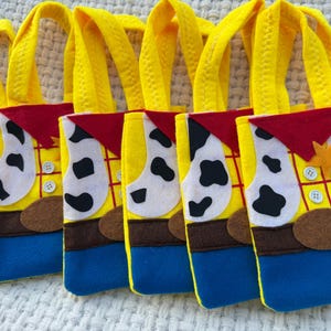 Puede incluir: Una fila de bolsas de mano de fieltro amarillas, azules, marrones, rojas y blancas. Cada bolsa est&aacute; dise&ntilde;ada para parecerse a un chaleco de vaquero con una insignia de sheriff, botones y una bandana roja. Las bolsas tienen asas amarillas.