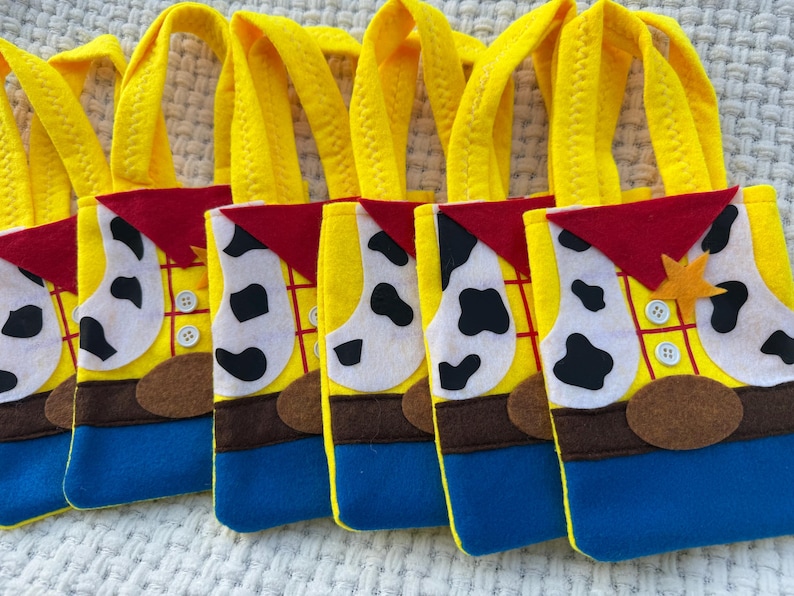 Puede incluir: Una fila de bolsas de mano de fieltro amarillas, azules, marrones, rojas y blancas. Cada bolsa est&aacute; dise&ntilde;ada para parecerse a un chaleco de vaquero con una insignia de sheriff, botones y una bandana roja. Las bolsas tienen asas amarillas.
