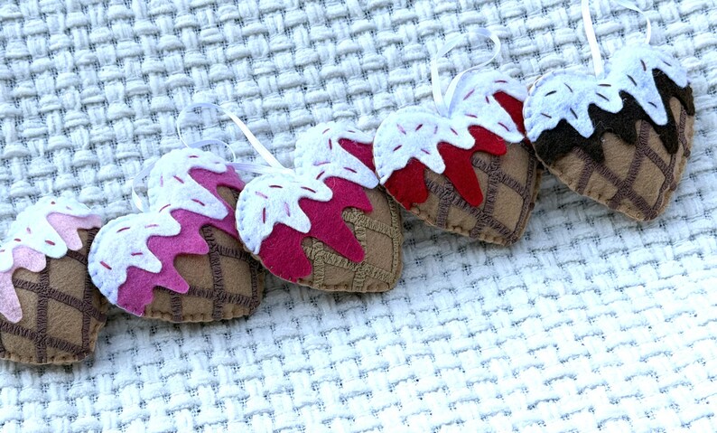 May include: A collection of heart-shaped ornaments resembling ice cream cones. Each ornament features a brown cone with a waffle pattern, topped with white "ice cream" and colorful sprinkles. The "ice cream" comes in shades of pink, red, and brown.