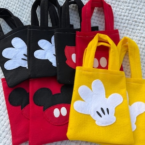 Może przedstawiać: Kolekcja małych torebek w kolorze czarnym, czerwonym i ż&oacute;łtym. Każda torba zawiera element projektu znanej postaci z kresk&oacute;wek, w tym białe rękawiczki, czerwoną muszkę i czarne uszy. Torby mają pasujące uchwyty.