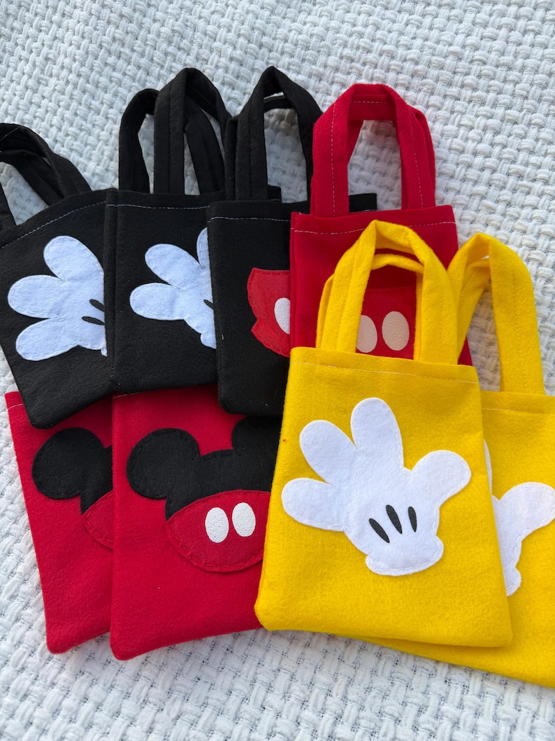 Może przedstawiać: Kolekcja małych torebek w kolorze czarnym, czerwonym i ż&oacute;łtym. Każda torba zawiera element projektu znanej postaci z kresk&oacute;wek, w tym białe rękawiczki, czerwoną muszkę i czarne uszy. Torby mają pasujące uchwyty.