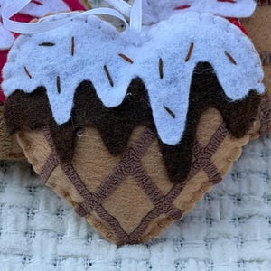 May include: Heart-shaped felt ornament resembling an ice cream cone. The ornament features a brown waffle cone base, chocolate sauce, and white frosting with brown sprinkles. A white ribbon is attached for hanging.