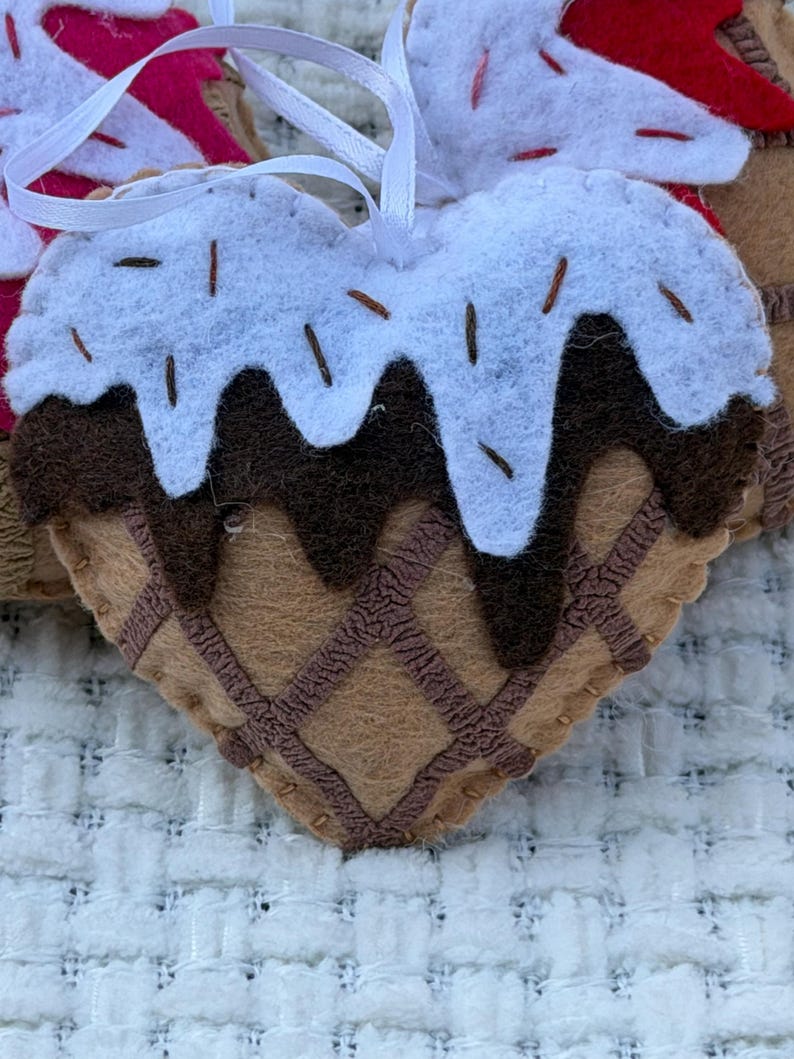 May include: Heart-shaped felt ornament resembling an ice cream cone. The ornament features a brown waffle cone base, chocolate sauce, and white frosting with brown sprinkles. A white ribbon is attached for hanging.
