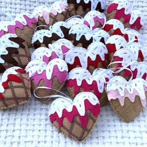 May include: A collection of heart-shaped felt ornaments resembling ice cream cones. The ornaments feature brown, pink, and white colors, with a waffle cone design and white ribbon for hanging. The ornaments are arranged on a textured white surface.