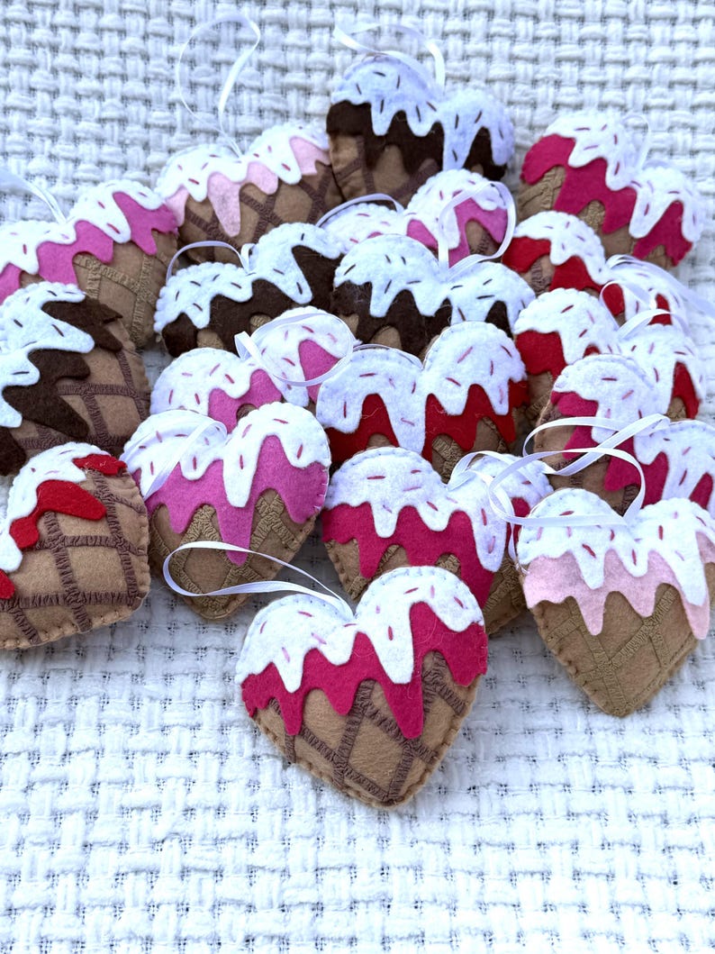 May include: A collection of heart-shaped felt ornaments resembling ice cream cones. The ornaments feature brown, pink, and white colors, with a waffle cone design and white ribbon for hanging. The ornaments are arranged on a textured white surface.