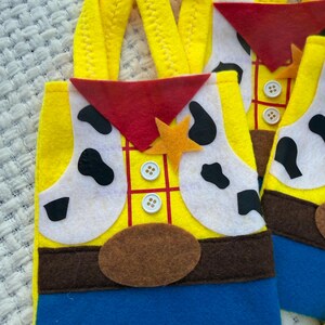 Puede incluir: Bolsas de mano de fieltro amarillo con un dise&ntilde;o inspirado en un personaje de vaquero. Las bolsas presentan un chaleco amarillo con parches blancos, un pa&ntilde;uelo rojo y un cintur&oacute;n marr&oacute;n. Las asas son amarillas y la parte inferior es azul.