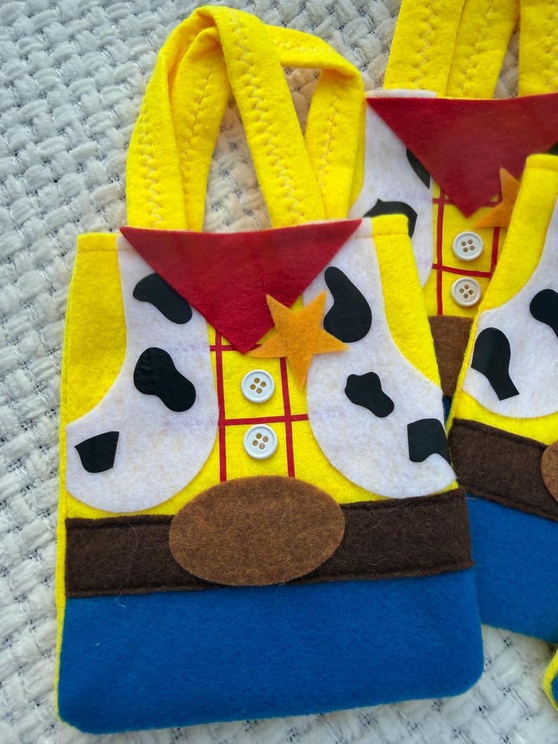 Puede incluir: Bolsas de mano de fieltro amarillo con un dise&ntilde;o inspirado en un personaje de vaquero. Las bolsas presentan un chaleco amarillo con parches blancos, un pa&ntilde;uelo rojo y un cintur&oacute;n marr&oacute;n. Las asas son amarillas y la parte inferior es azul.