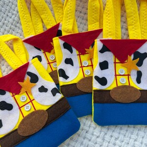 Puede incluir: Una colecci&oacute;n de bolsas de fieltro amarillas, azules, rojas y blancas. Las bolsas est&aacute;n dise&ntilde;adas para parecerse a un personaje de dibujos animados, con una camisa amarilla, pantalones azules, un pa&ntilde;uelo rojo y un cintur&oacute;n marr&oacute;n. Cada bolsa tiene un asa amarilla y una insignia en forma de estrella.