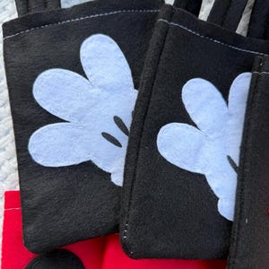 Może przedstawiać: Zbliżenie kilku czarnych filcowych toreb z białymi aplikacjami rękawic Myszki Miki. Torby mają czarne uchwyty i są zszyte białą nicią. Czerwony filc jest widoczny na dole toreb.