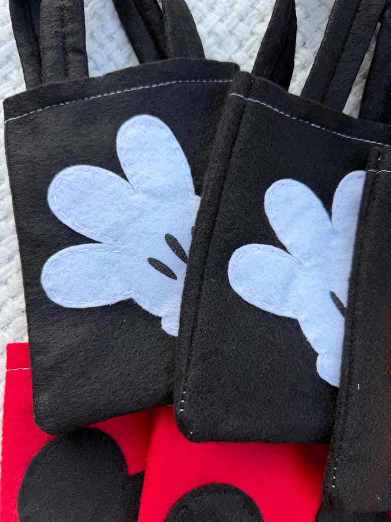 Może przedstawiać: Zbliżenie kilku czarnych filcowych toreb z białymi aplikacjami rękawic Myszki Miki. Torby mają czarne uchwyty i są zszyte białą nicią. Czerwony filc jest widoczny na dole toreb.