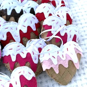 May include: A collection of heart-shaped felt ornaments resembling ice cream cones. The ornaments feature a brown cone base, a pink or red ice cream layer, white frosting, and red sprinkles. Each ornament has a white ribbon for hanging.