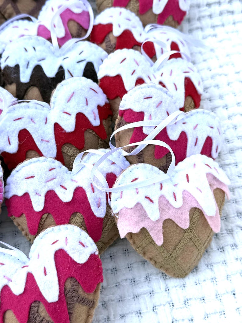 May include: A collection of heart-shaped felt ornaments resembling ice cream cones. The ornaments feature a brown cone base, a pink or red ice cream layer, white frosting, and red sprinkles. Each ornament has a white ribbon for hanging.