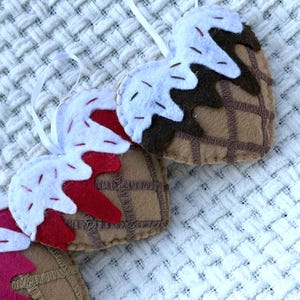 May include: Handmade felt ice cream cone ornaments in a heart shape. The cones are brown with a waffle pattern, topped with white and red felt "ice cream" and sprinkles. Each ornament has a white ribbon for hanging.