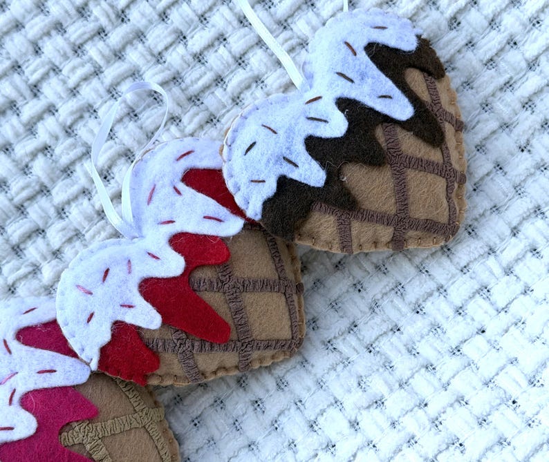 May include: Handmade felt ice cream cone ornaments in a heart shape. The cones are brown with a waffle pattern, topped with white and red felt "ice cream" and sprinkles. Each ornament has a white ribbon for hanging.