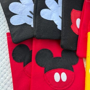 Może przedstawiać: Kolekcja filcowych torebek w kolorach czarnym, czerwonym i ż&oacute;łtym. Czarne torebki mają białe rękawice Myszki Miki, a czerwone torebki przedstawiają twarz Myszki Miki z białymi oczami. Widoczna jest r&oacute;wnież ż&oacute;łta torebka.