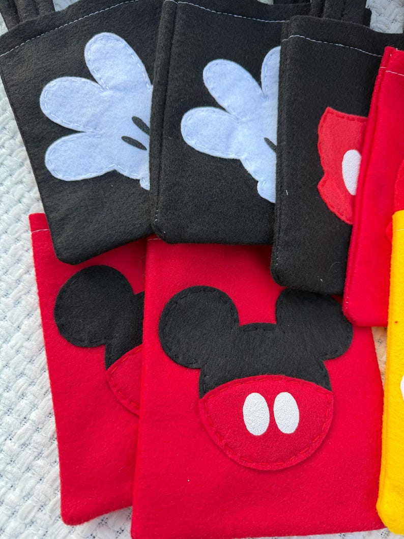 Może przedstawiać: Kolekcja filcowych torebek w kolorach czarnym, czerwonym i ż&oacute;łtym. Czarne torebki mają białe rękawice Myszki Miki, a czerwone torebki przedstawiają twarz Myszki Miki z białymi oczami. Widoczna jest r&oacute;wnież ż&oacute;łta torebka.