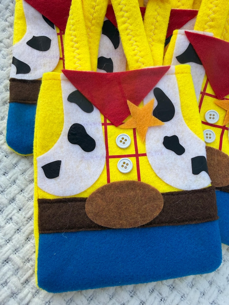 Puede incluir: Bolsas de fieltro amarillas con un dise&ntilde;o inspirado en un personaje de vaquero. Las bolsas presentan un pa&ntilde;uelo rojo, detalles blancos con manchas negras, cintur&oacute;n marr&oacute;n y una insignia en forma de estrella. Se utiliza fieltro azul y amarillo para la base de las bolsas.