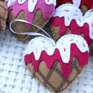 May include: Heart-shaped felt ornaments with a whimsical ice cream cone design. The ornaments feature a brown waffle cone base, pink ice cream, white frosting, and red sprinkles. Each ornament has a white ribbon for hanging.