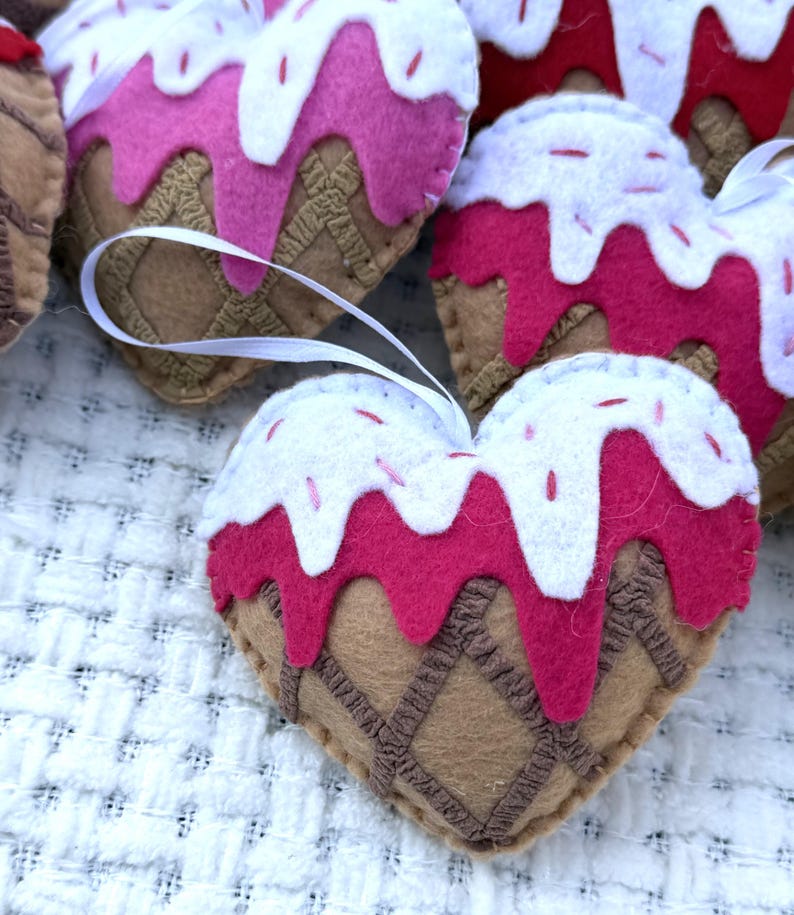 May include: Heart-shaped felt ornaments with a whimsical ice cream cone design. The ornaments feature a brown waffle cone base, pink ice cream, white frosting, and red sprinkles. Each ornament has a white ribbon for hanging.