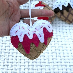 May include: A felt heart-shaped ornament resembling an ice cream cone. The ornament features a brown waffle cone base, a pink ice cream layer, and white frosting with red sprinkles. A white ribbon is attached for hanging.