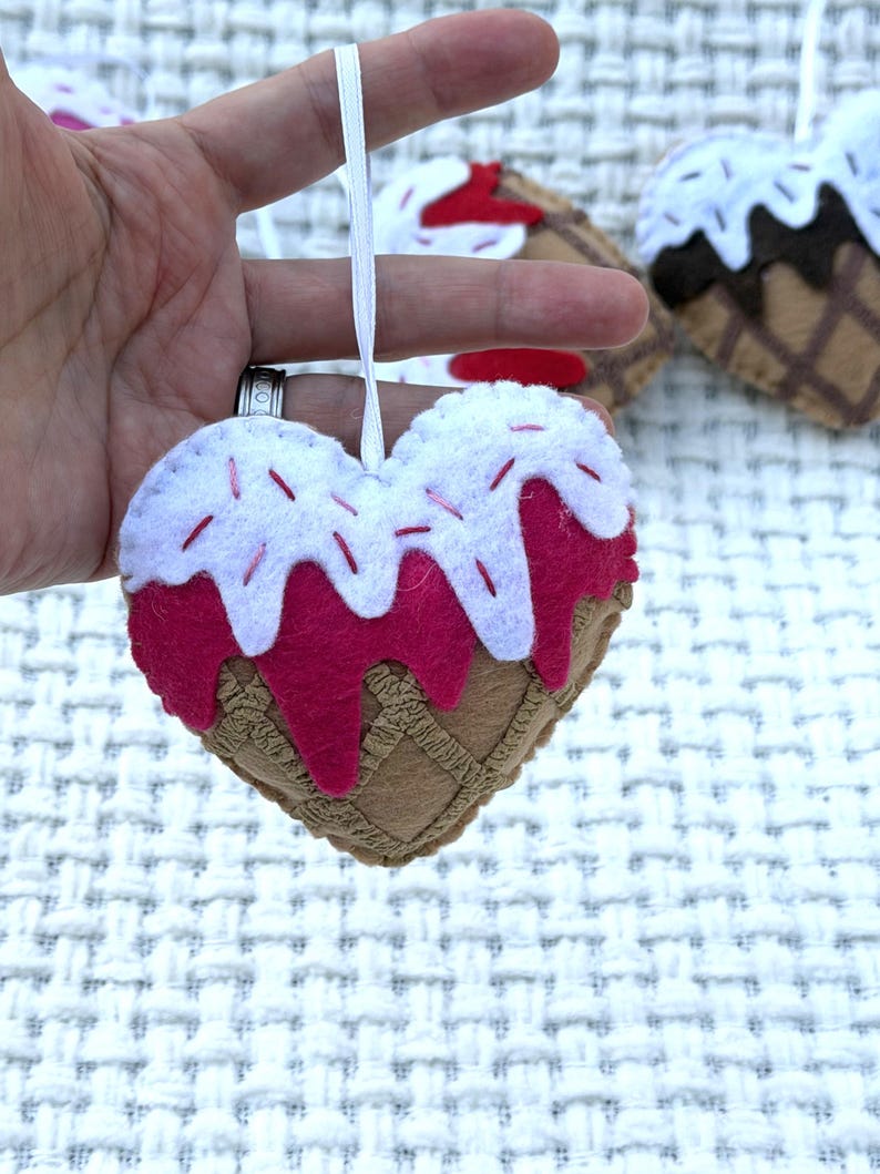 May include: A felt heart-shaped ornament resembling an ice cream cone. The ornament features a brown waffle cone base, a pink ice cream layer, and white frosting with red sprinkles. A white ribbon is attached for hanging.