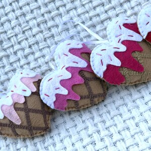 May include: A close-up of several heart-shaped felt ornaments resembling ice cream cones. Each ornament features a brown waffle cone base, topped with pink and white felt "ice cream" and white ribbon for hanging. The ornaments are arranged on a white textured surface.