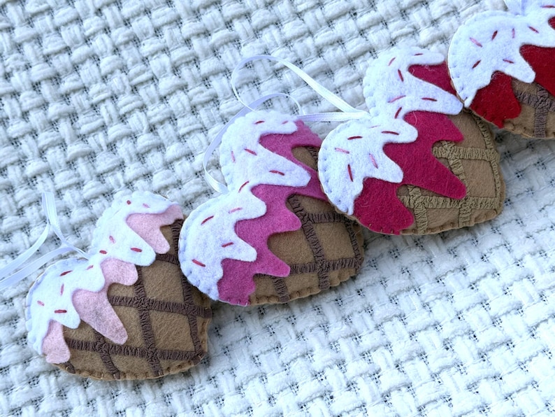 May include: A close-up of several heart-shaped felt ornaments resembling ice cream cones. Each ornament features a brown waffle cone base, topped with pink and white felt "ice cream" and white ribbon for hanging. The ornaments are arranged on a white textured surface.
