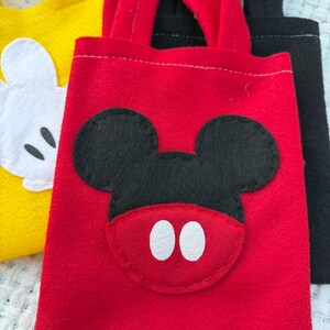 Może przedstawiać: Czerwona filcowa torba z czarną aplikacją z twarzą Myszki Miki i białymi owalnymi oczami. Ż&oacute;łta torba z białą twarzą Myszki Miki jest widoczna po lewej stronie. Czarna torba znajduje się po prawej stronie.