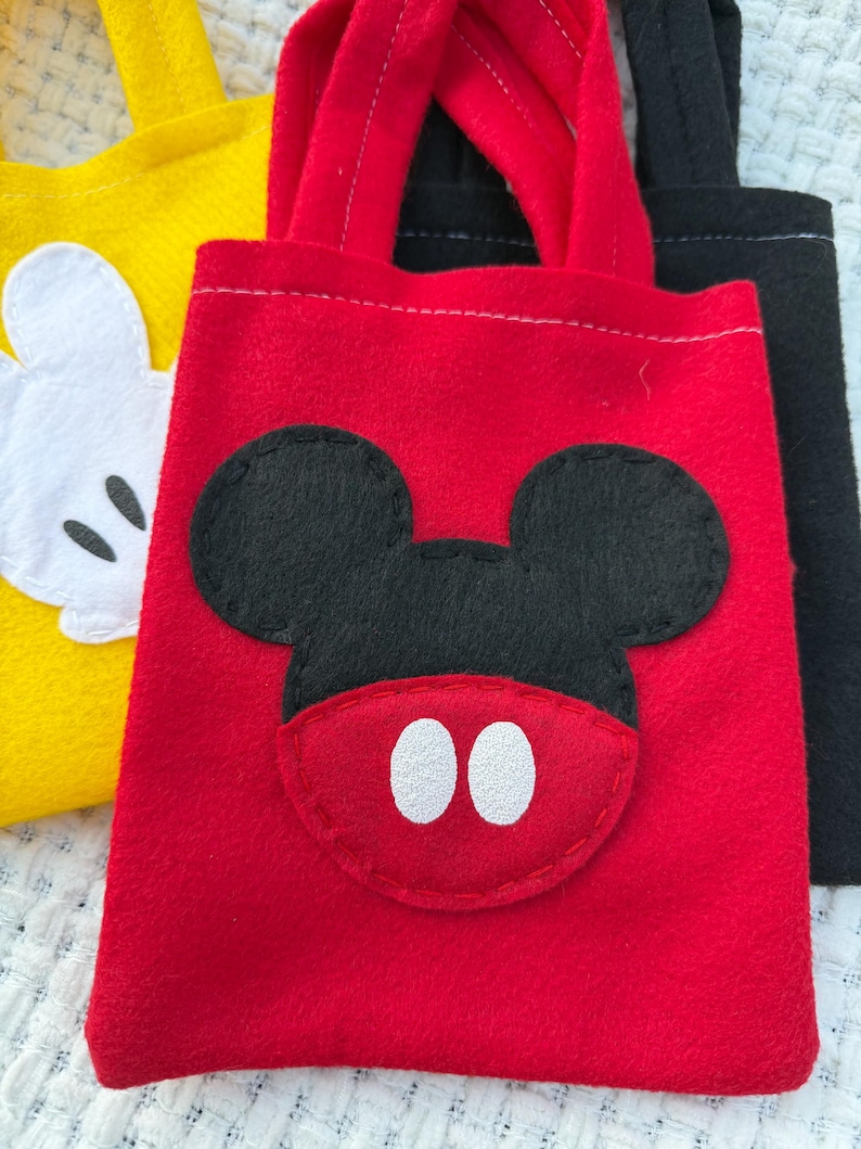 Może przedstawiać: Czerwona filcowa torba z czarną aplikacją z twarzą Myszki Miki i białymi owalnymi oczami. Ż&oacute;łta torba z białą twarzą Myszki Miki jest widoczna po lewej stronie. Czarna torba znajduje się po prawej stronie.