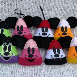 May include: A collection of handmade felt ornaments in the shape of candy corn with Mickey and Minnie Mouse faces. The ornaments feature black mouse ears and are decorated with various colors, including green, pink, red, orange, and white.