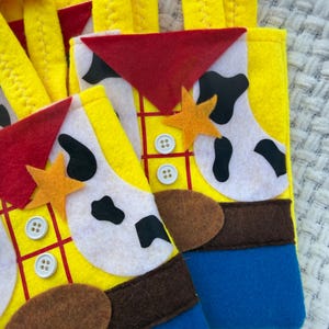 Puede incluir: Calcetines navide&ntilde;os de fieltro amarillo con tem&aacute;tica de Woody de Toy Story. Los calcetines tienen un tri&aacute;ngulo rojo, estampado de vaca blanco y negro, detalles de fieltro marr&oacute;n y fondos de fieltro azul. Cada calcet&iacute;n tiene una estrella y detalles de botones.