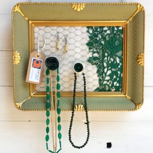 Può includere: Un organizer da parete rettangolare per gioielli con cornice verde salvia e oro, sfondo in rete metallica bianca e un pannello in pizzo verde. Include due pomelli che sostengono collane di perline verdi e un cartellino in carta marrone.