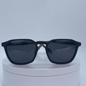 May include: Black sunglasses with square frames and dark lenses. The sunglasses have a matte black finish on the front and temples with a light brown interior. The sunglasses are displayed on a white surface.