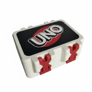 Peut inclure: Un étui de jeu de cartes UNO en plastique blanc avec des fermoirs rouges. Le couvercle présente un fond noir avec le mot "UNO" en lettres blanches à l'intérieur d'un ovale rouge. L'étui est rectangulaire et conçu pour contenir un jeu de cartes UNO.