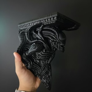 Puede incluir: Un soporte de estante negro y detallado que representa un Xenomorfo Alien. El soporte tiene una parte superior plana y un diseño esculpido y texturizado. Se sostiene en una mano sobre un fondo oscuro.