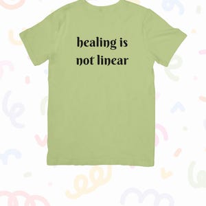 以下が含まれることがあります： 「healing is not linear」の黒い文字がプリントされた、薄い緑色のTシャツ。Tシャツは丸首で半袖です。スローガンは、衣服の前面中央に印刷されています。