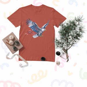 Könnte beinhalten: Ein rostfarbenes T-Shirt mit einer detaillierten Illustration eines Weißkopfseeadlers im Flug. Der Adler hat weiße und blaue Federn und hält ein rotes Band. Der Text auf dem Bild lautet "Digital Download Only | High-Res Transparent PNG | No Physical Item Shipped | Personal Use Only".