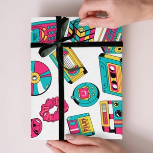 Peut inclure: Papier cadeau blanc avec un ruban noir, orné d'un motif coloré d'appareils électroniques rétro, tels que des cassettes, des CD et un Rubik's Cube. Le texte sur l'emballage dit "FILES".