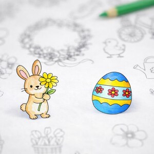 Puede incluir: Ilustración coloreada de un conejito de Pascua sosteniendo una flor amarilla junto a un huevo de Pascua decorado. El huevo es azul con rayas amarillas y acentos de flores rojas. Un lápiz verde está en el fondo.
