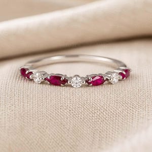 Op de afbeelding: Een zilverkleurige ring met een afwisselend patroon van dieprode stenen in marquise-slijpvorm en heldere witte stenen in ronde slijpvorm, gezet langs de band. De metalen ring is dun en gepolijst en toont een symmetrische opstelling.