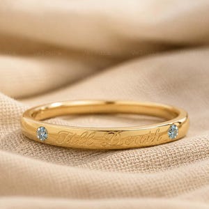 Könnte beinhalten: Ein goldener Ring mit der eingravierten Schrift Till Death in Schreibschrift, akzentuiert durch zwei kleine, runde, hellblaue Edelsteine, die auf beiden Seiten der Inschrift in das Metall eingelassen sind und einen polierten Glanz aufweisen.