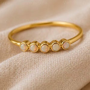 以下が含まれることがあります： 細く滑らかなバンドを備えたゴールドトーンのリング。リングの上部には、個別の円形ベゼルにセットされた、オパールのような5つの小さな丸い虹色の白い石が水平に並んでいます。