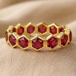 Könnte beinhalten: Ein goldener Ring mit einer durchgehenden Reihe von sechseckig geschliffenen roten Edelsteinen. Jeder Stein ist in eine sechseckige Goldfassung eingefasst, wodurch ein wabenartiges Muster entsteht, das den gesamten Ring umschließt.