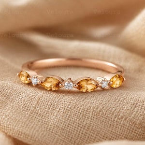 Op de afbeelding: Roségouden ring met afwisselend marquise-geslepen gele edelstenen en kleine ronde witte edelstenen. De ring is geplaatst op een neutraal gekleurde stoffen achtergrond. Een delicaat sieraad.