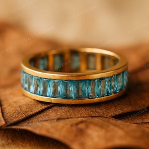 May include: A gold band ring featuring a row of rectangular, light blue gemstones. The ring is set against a brown, textured background, creating a contrast between the warm gold and cool blue tones. The ring's design is simple yet elegant.