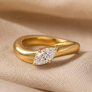 Puede incluir: Un anillo de oro pulido con una piedra preciosa transparente de talla marquesa engastada horizontalmente en el centro. La banda tiene un perfil liso, grueso y redondeado que se estrecha ligeramente hacia el engaste, creando un aspecto fluido.