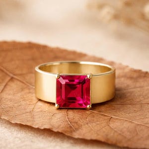 Könnte beinhalten: Ein Goldring mit einem quadratisch geschliffenen, leuchtend roten Edelstein in der Mitte. Der Ring hat eine breite, polierte Metallschiene, die den facettierten roten Stein mit vier kleinen Krappen sicher hält und ein klares Design bildet.