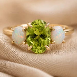 May include: A gold ring featuring a large oval-cut peridot gemstone flanked by two smaller opal gemstones. The peridot is a vibrant green, while the opals have a milky white appearance. The ring is set on a gold band.